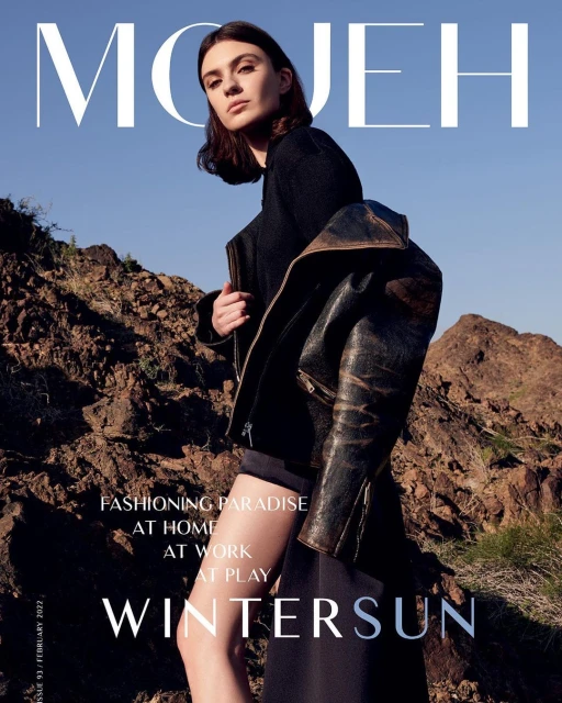 Fashion magazine cover featuring a female model wearing a black jacket and dark skirt, posing against a backdrop of rocky terrain under a blue sky.
