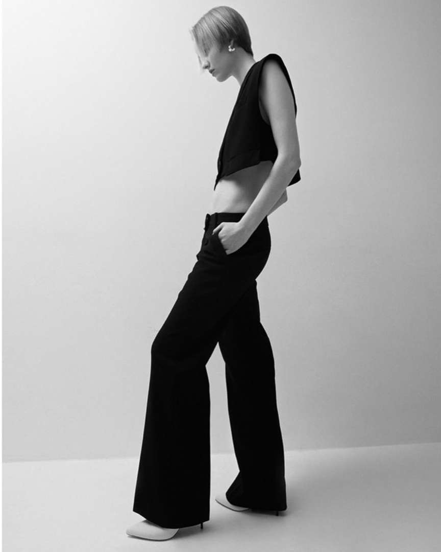 Fashionable adult female with short blonde hair poses in a cropped vest and wide-leg black pants against a pale neutral background.