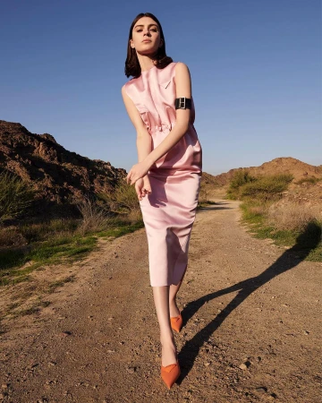 A young woman in a light pink dress and orange heels poses on a desert trail with mountains and a blue sky in the background.