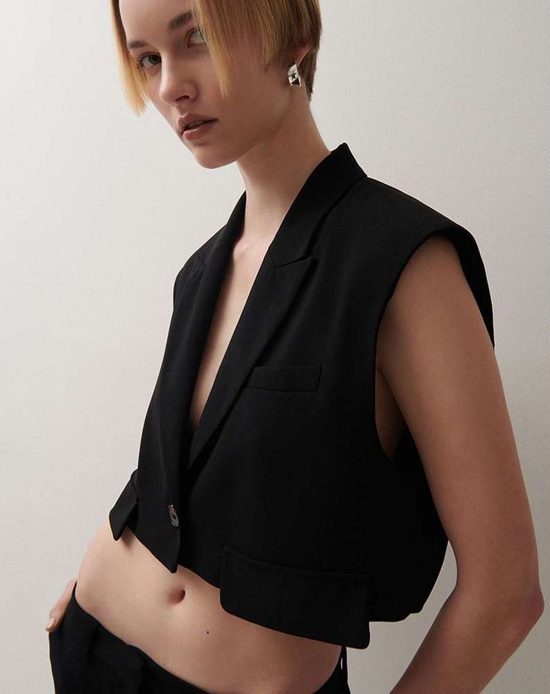 Fashionable adult woman posing in a black cropped vest and pants against a neutral backdrop in a studio setting, minimal styling.