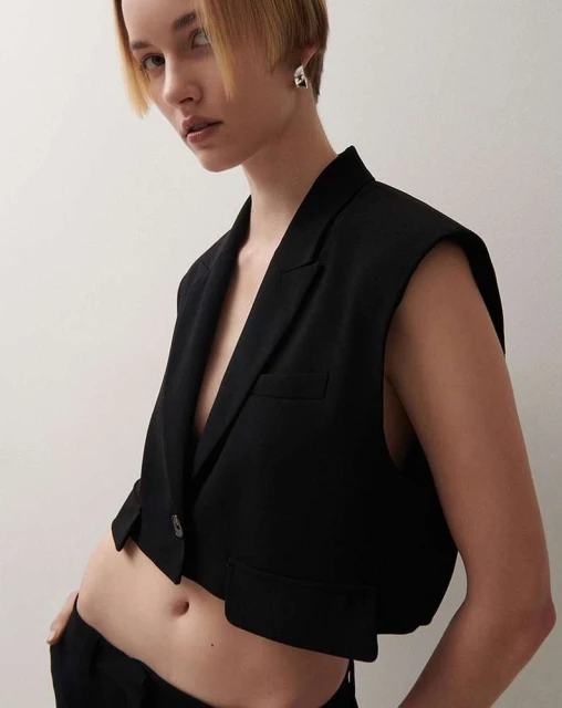 Fashionable adult woman posing in a black cropped vest and pants against a neutral backdrop in a studio setting, minimal styling.