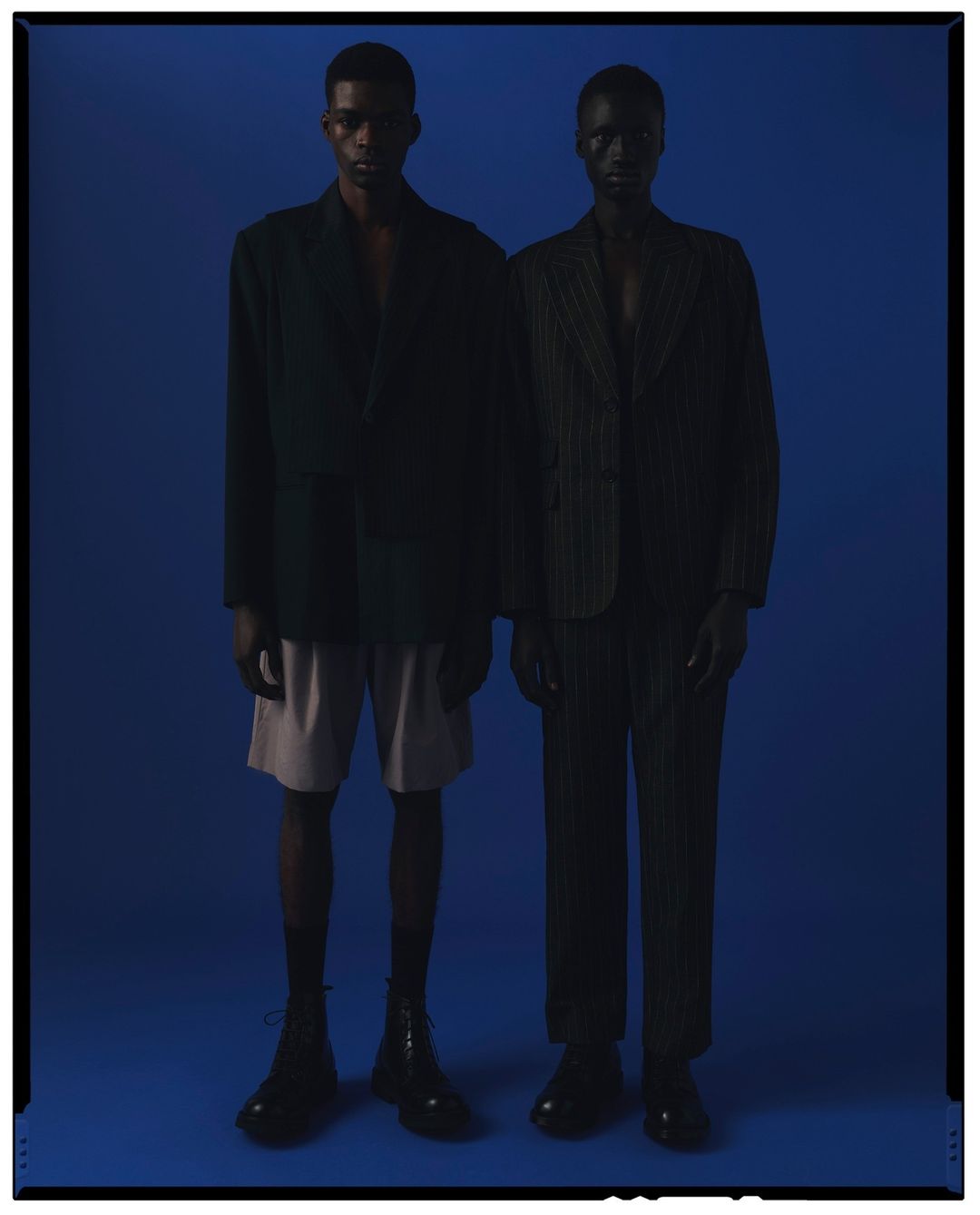 Two dark-skinned fashion models in blazers, one with shorts, stand in a blue photo studio with subdued, shadowy lighting.