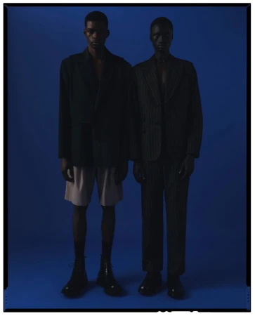 Two dark-skinned fashion models in blazers, one with shorts, stand in a blue photo studio with subdued, shadowy lighting.