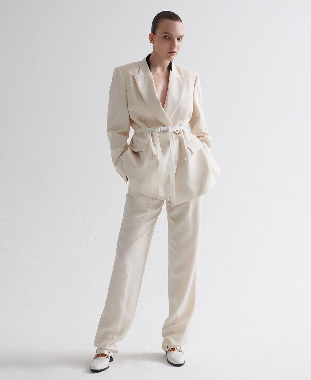Woman in an ivory-colored suit with a belt, standing in front of a white background in a fashion studio. Full shot of the outfit.