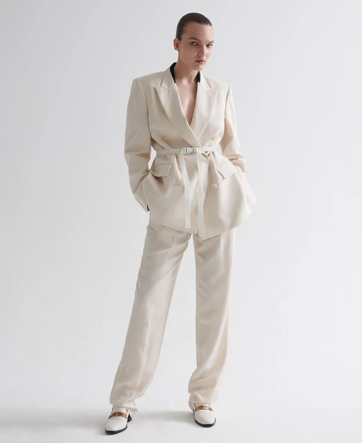Woman in an ivory-colored suit with a belt, standing in front of a white background in a fashion studio. Full shot of the outfit.