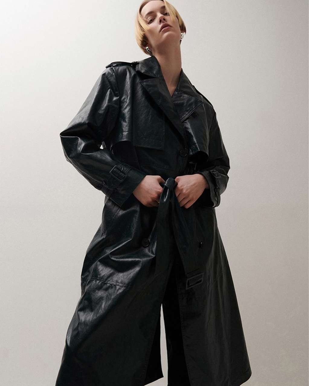 Model wearing a long black leather trench coat against a plain light background