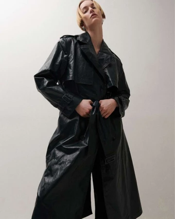Model wearing a long black leather trench coat against a plain light background