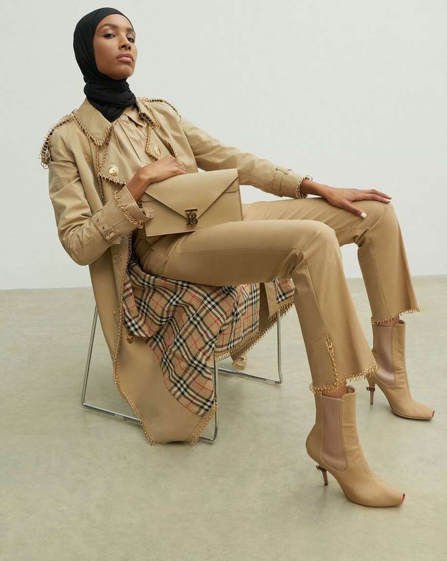 Adult female seated in a beige trench coat, pants, and boots, holding a handbag, in a studio shot.