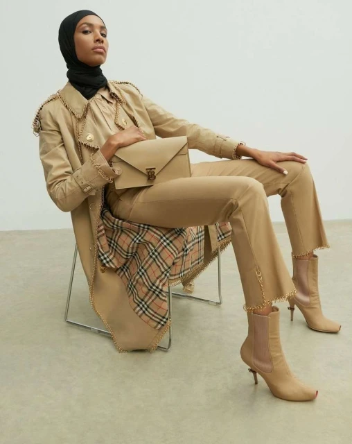 Fashionable woman in trench coat, hijab, trousers, and ankle boots, seated in a studio with a neutral-toned handbag and plaid accent.