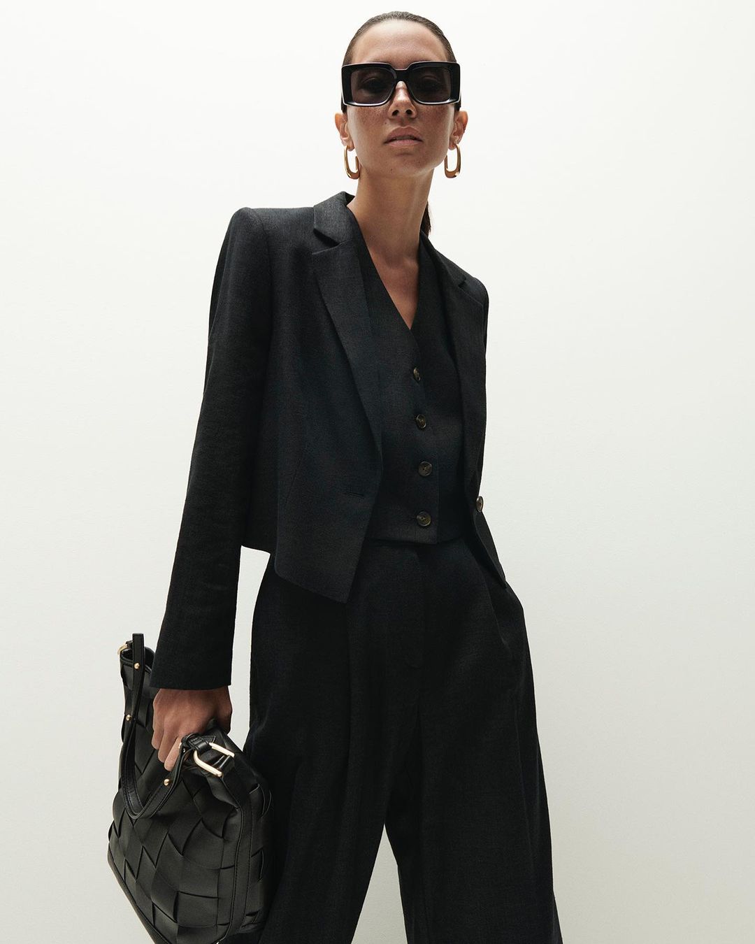 Model in black suit wearing sunglasses and holding a woven bag against a bright white background