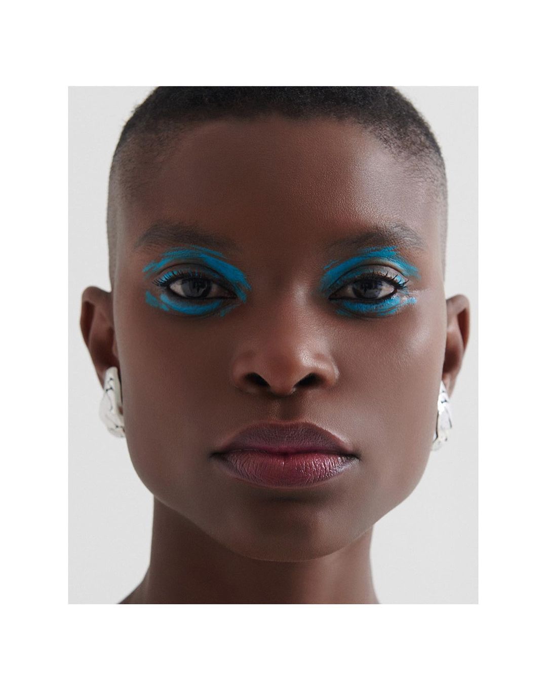 Close-up portrait of a Black woman with shaved head and dramatic teal eye makeup, wearing geometric silver earrings against a grey background.