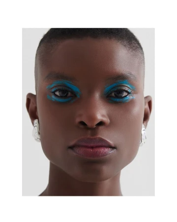 Close-up portrait of a Black woman with shaved head and dramatic teal eye makeup, wearing geometric silver earrings against a grey background.