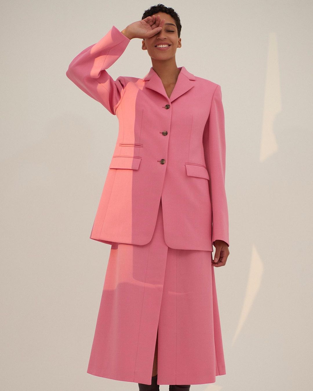 A smiling adult woman in a pink suit with a blazer and skirt poses in front of a neutral background with soft, natural lighting.