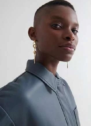 Close-up of a dark-skinned adult female model with an undercut wearing gold dangling earrings and a blue collared shirt against a white background.