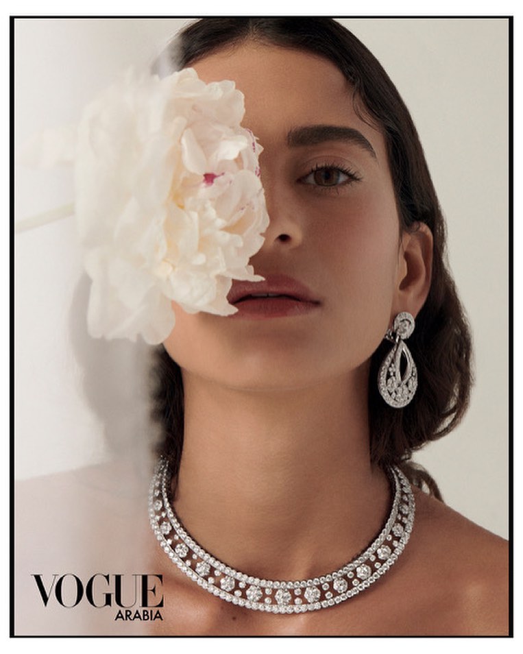 Close-up portrait of a woman holding a large white flower over her eye, wearing a diamond necklace and earrings.