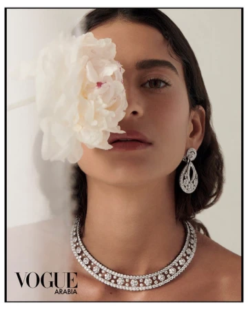 Close-up portrait of a woman holding a large white flower over her eye, wearing a diamond necklace and earrings.