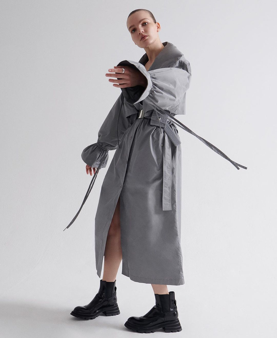 Adult female model in studio wearing a grey trench coat with large sleeves and black boots. Fashion shot with grey and white background.