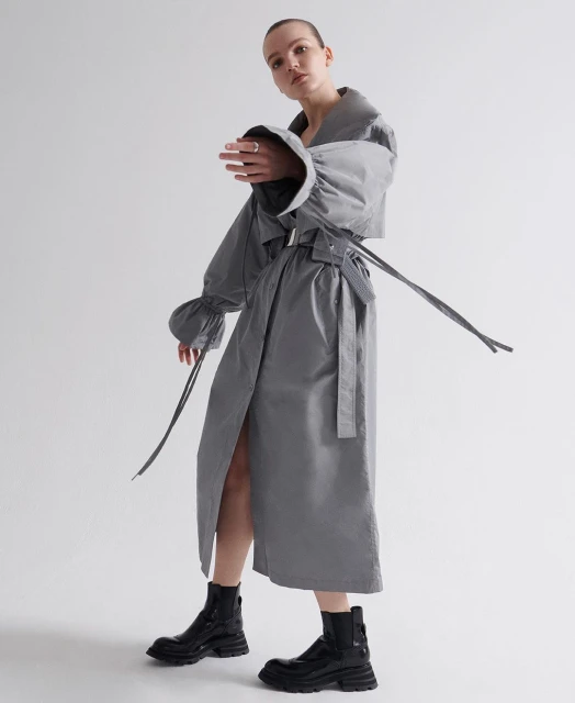 Adult female model in studio wearing a grey trench coat with large sleeves and black boots. Fashion shot with grey and white background.