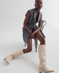 Model with short hair wearing a grey sleeveless vest dress and tall cream boots seated on a metal chair against a white background.