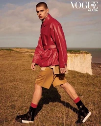 Young male model in a red leather jacket and beige shorts walking outdoors on a coastal cliffside by Louis Christopher.