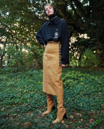 Model wearing a black shirt, plaid skirt, and matching boots standing in a lush forest by Louis Christopher