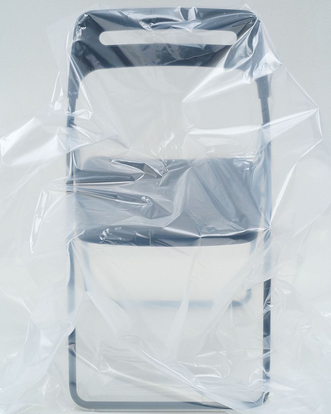 Still life of a dark grey folding chair partially covered in a clear plastic sheet, studio lit against a white background.