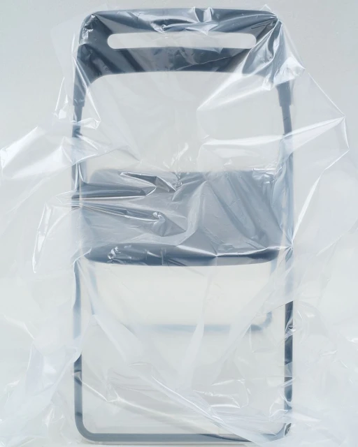 Still life of a dark grey folding chair partially covered in a clear plastic sheet, studio lit against a white background.