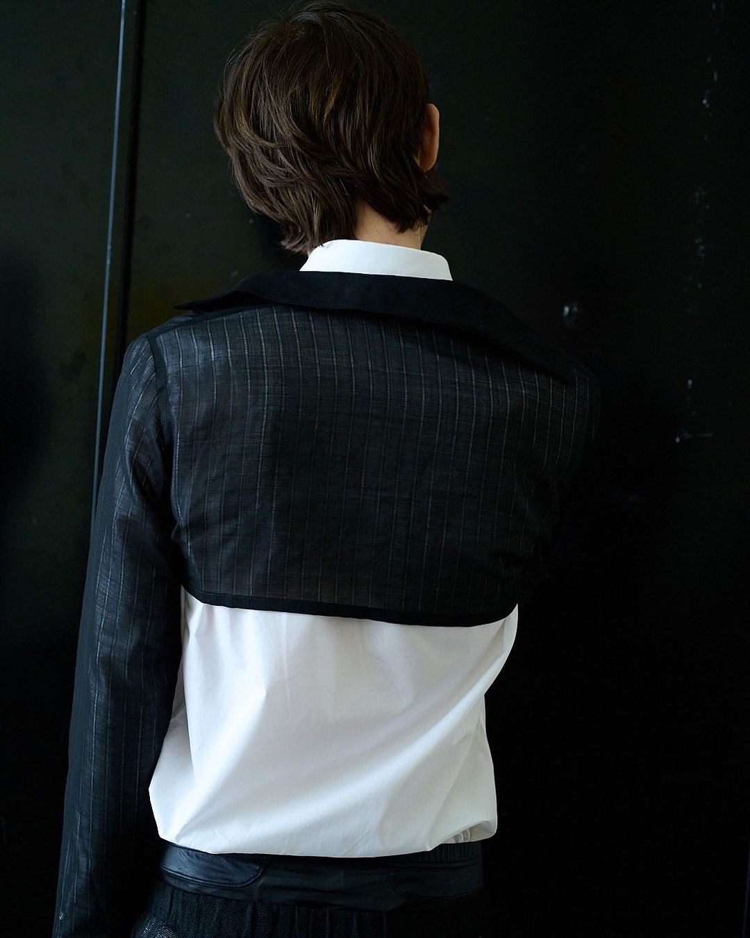 Back view of a person in a black and white outfit standing against a black background in an indoor environment, fashion photography.
