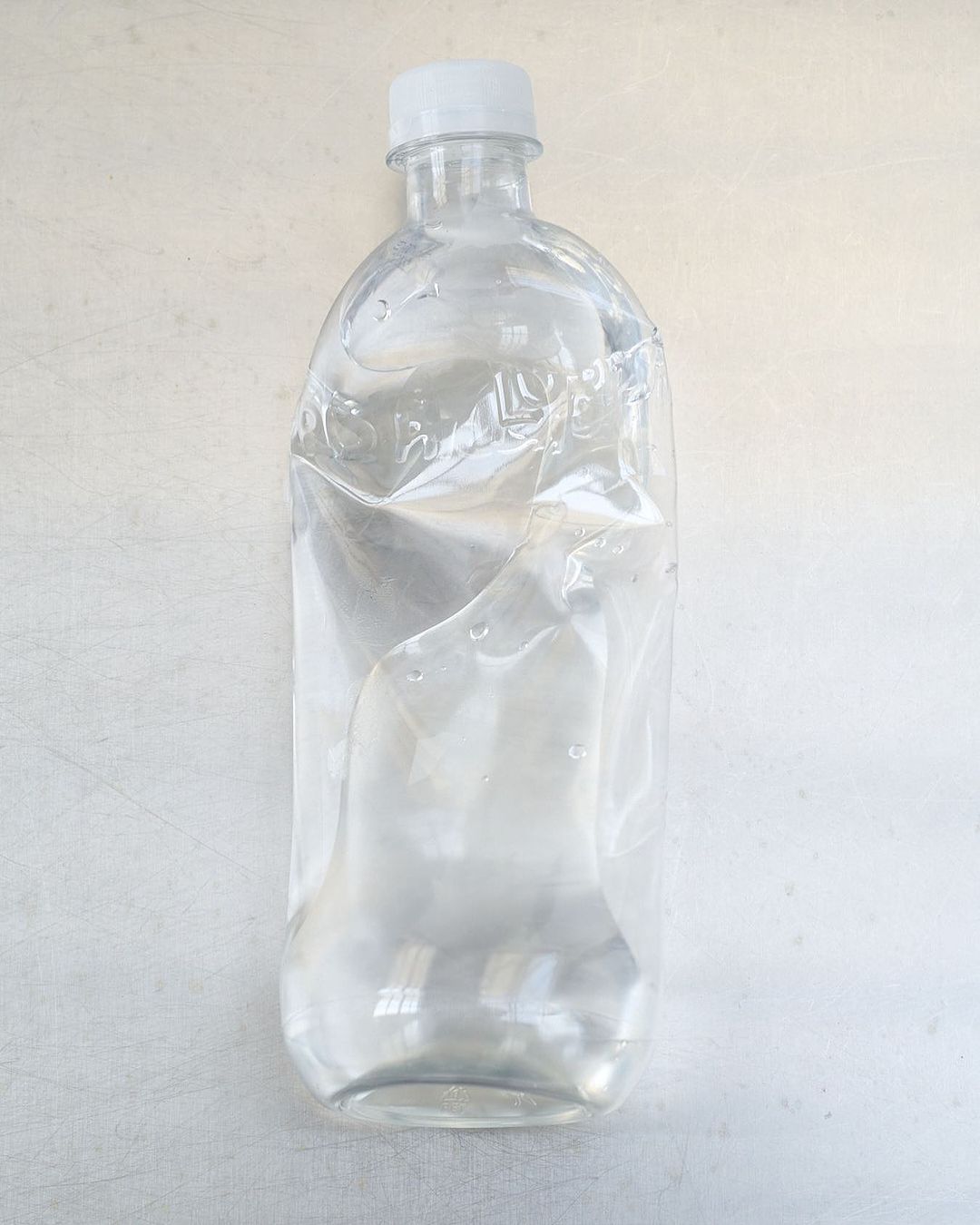 Clear, slightly crumpled water bottle on a white surface, with a white cap. The bottle is partially filled with water, minimal aesthetic.