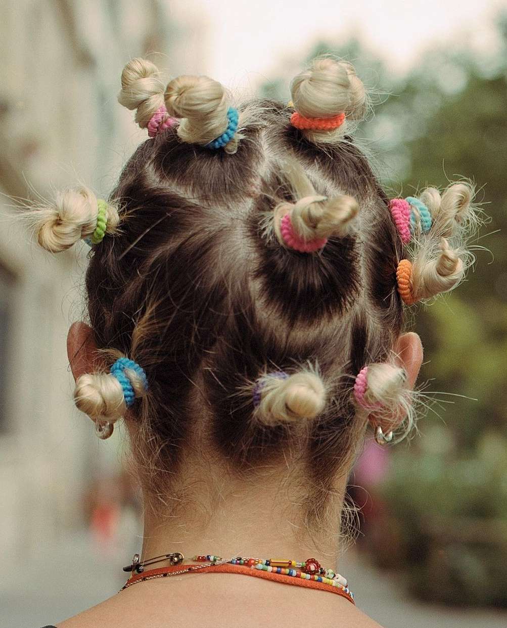 Close up from the back of a woman's head, her blonde hair styled in small knots held by colorful elastic bands outdoors.