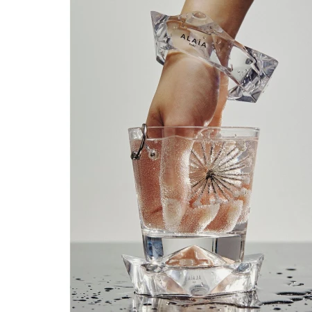 Clear Alaïa bracelet adorns a hand emerging from a glass of water; an identical bracelet serves as the glass's base, creating a minimalist beauty shot.