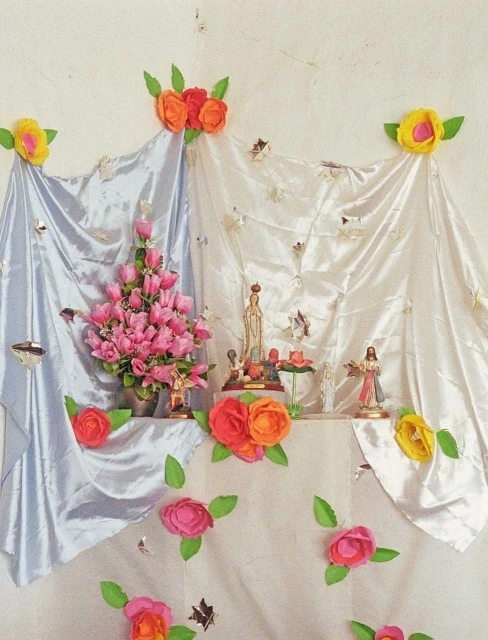 Religious altar with statues of Mary and Jesus adorned with pink and orange flowers and draped with silver and white satin cloths.