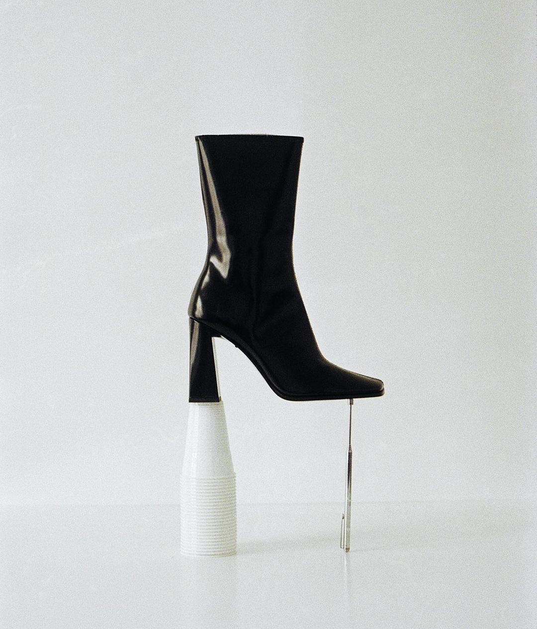 Black leather heeled boot artfully balanced on a white plastic cup with a chrome rod providing extra support in a clean, bright studio setting.