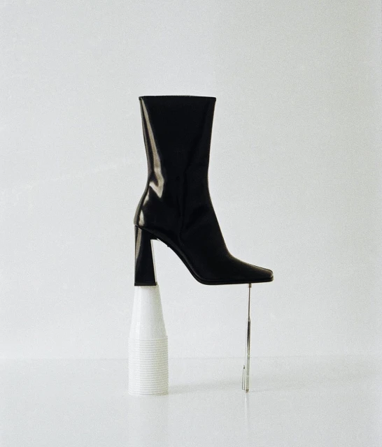 Black leather heeled boot artfully balanced on a white plastic cup with a chrome rod providing extra support in a clean, bright studio setting.