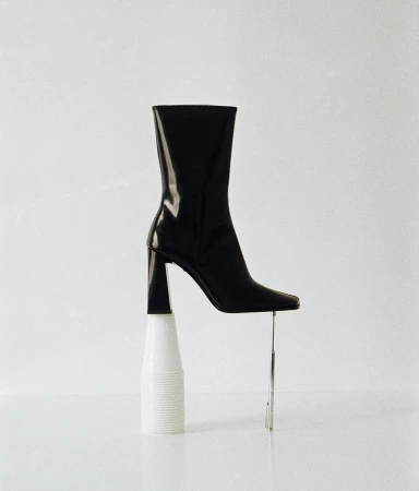 Black leather heeled boot artfully balanced on a white plastic cup with a chrome rod providing extra support in a clean, bright studio setting.