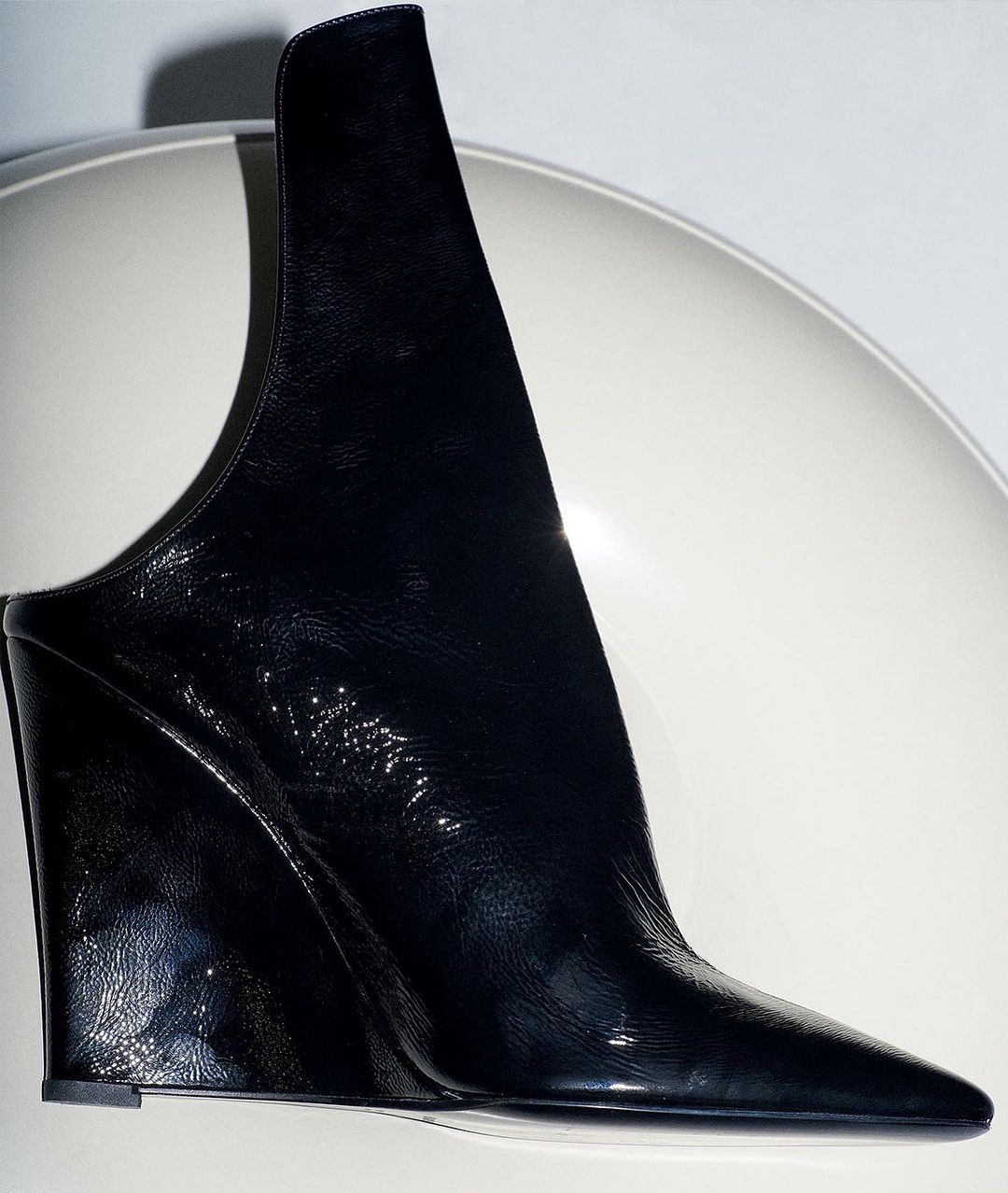 Close-up of a shiny, black patent leather high-fashion mule shoe with a wedge heel and a unique architectural design on a white surface.