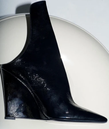 Close-up of a shiny, black patent leather high-fashion mule shoe with a wedge heel and a unique architectural design on a white surface.