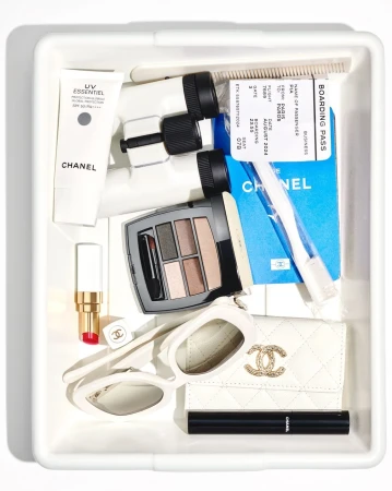 Overhead shot of a white tray containing Chanel beauty products, sunglasses, a wallet, and a boarding pass; all items are white.