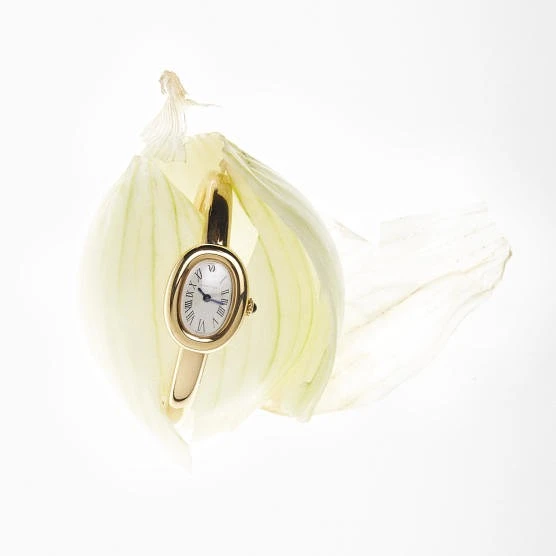 Elegant gold wristwatch with an oval face positioned within translucent onion layers, bathed in bright, even lighting against a white background.