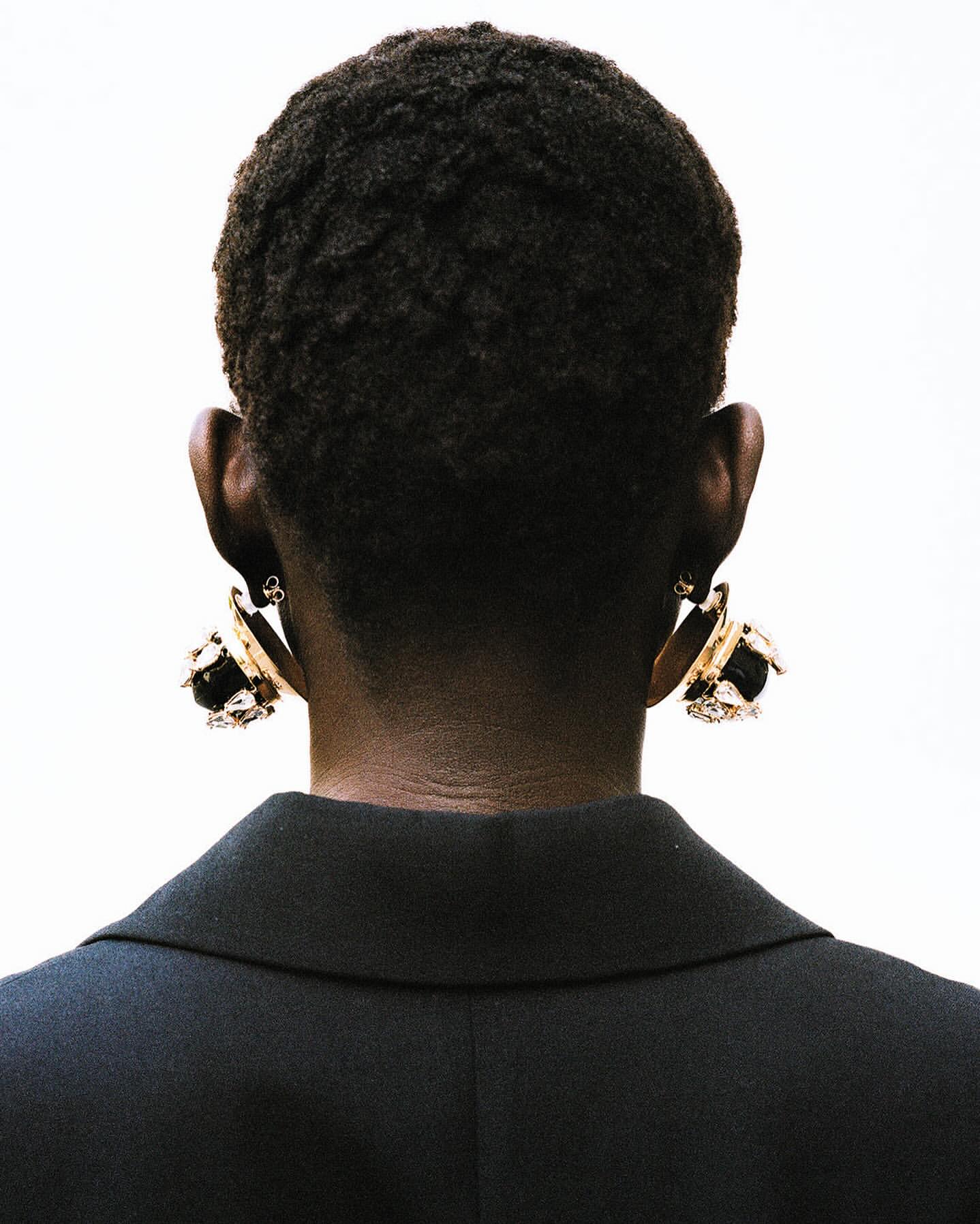 Back view of an adult female with dark skin and short, dark hair wearing gold earrings and a black blazer against a white backdrop.