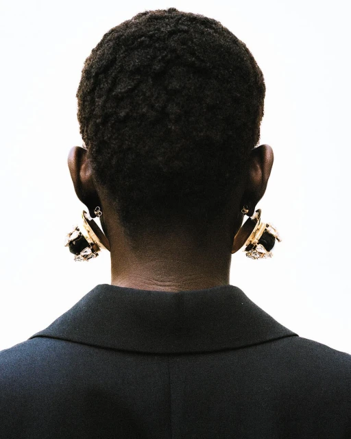 Back view of an adult female with dark skin and short, dark hair wearing gold earrings and a black blazer against a white backdrop.