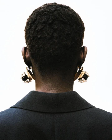 Back view of an adult female with dark skin and short, dark hair wearing gold earrings and a black blazer against a white backdrop.