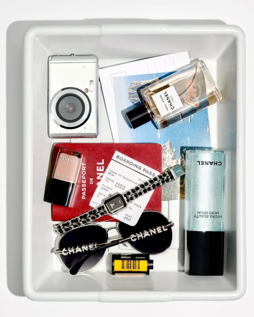 Overhead shot in a white tray filled with beauty and travel accessories: camera, passport, perfume, sunglasses, watch, and film.
