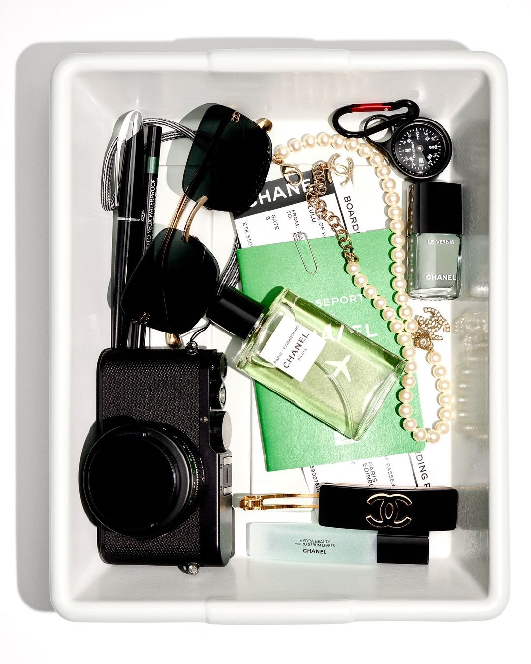 Overhead shot of a white tray containing a camera, passport, sunglasses, and Chanel beauty products, arranged in a flat lay.