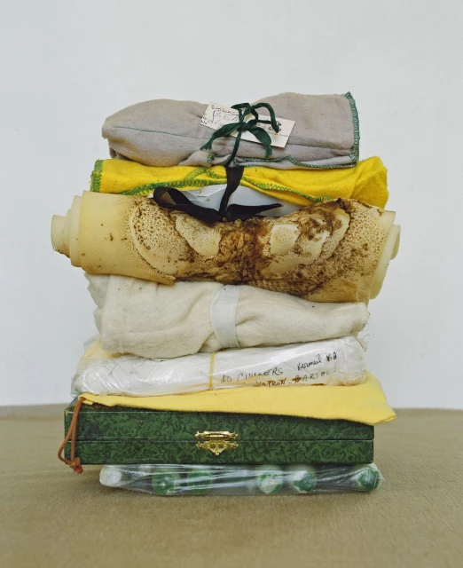A stack of wrapped objects rests on a table against a white background, with a dark green patterned box near the bottom and various fabrics.