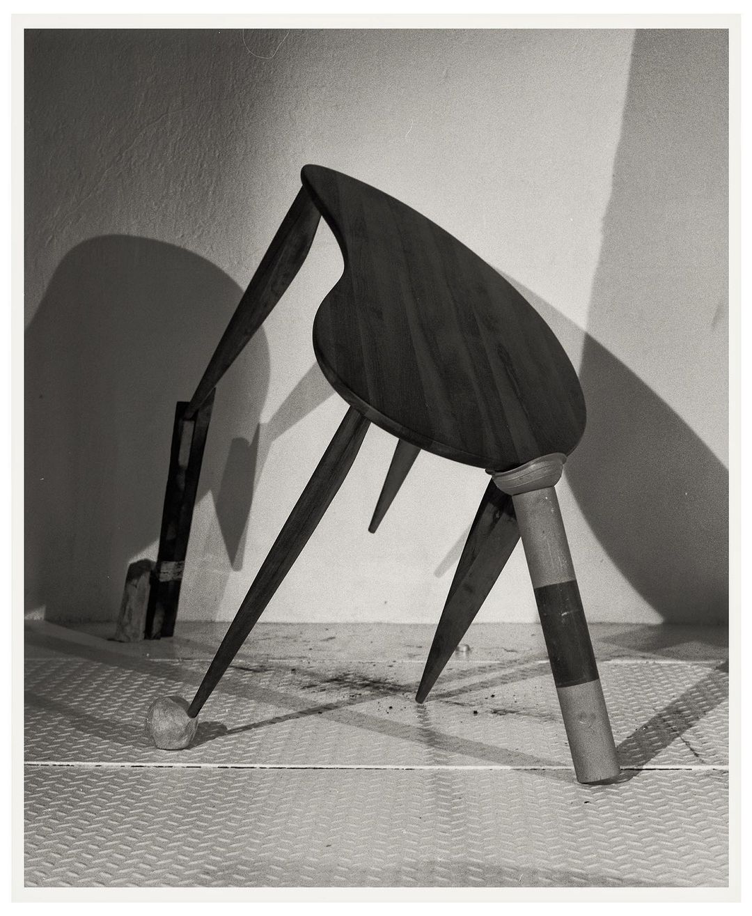 Monochromatic still life photograph of an altered wooden chair against a textured white wall in a studio setting, casting shadows.