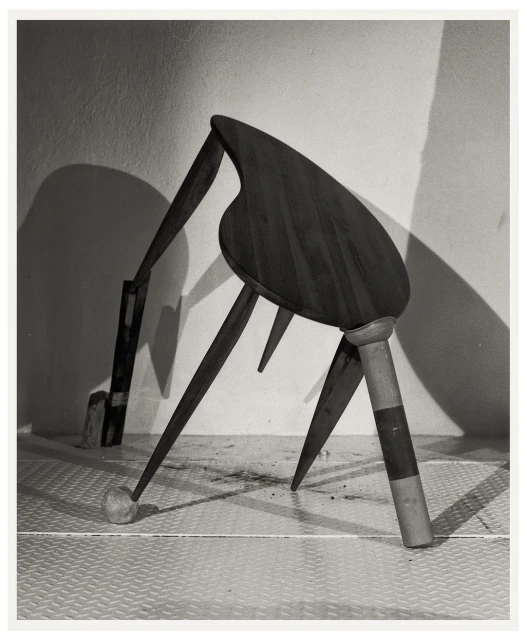 Monochromatic still life photograph of an altered wooden chair against a textured white wall in a studio setting, casting shadows.