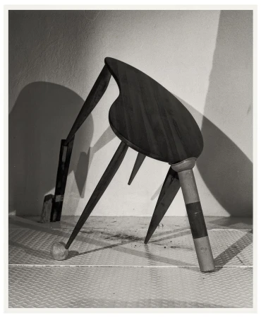 Monochromatic still life photograph of an altered wooden chair against a textured white wall in a studio setting, casting shadows.