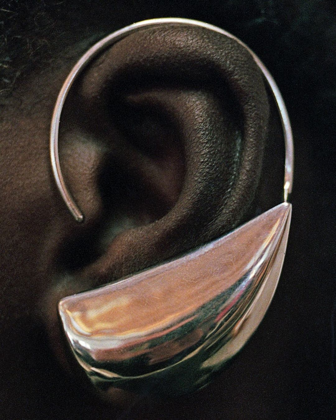 Close-up of an adult's ear wearing a sculptural, oversized silver earring with a reflective finish against a dark background.