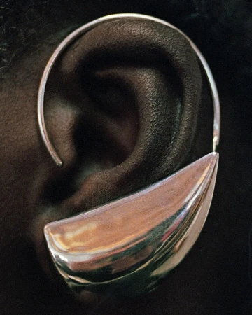 Close-up of an adult's ear wearing a sculptural, oversized silver earring with a reflective finish against a dark background.